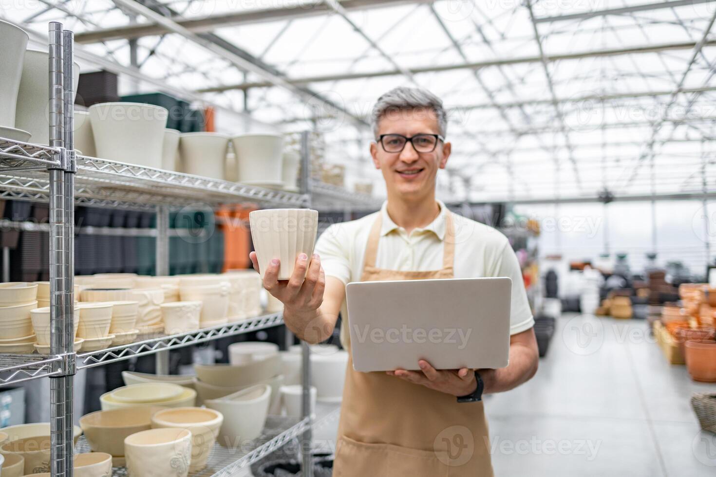 A shopkeeper holding a flowerpot and laptop, surrounded by various ceramic pots, in a plant shop. The man is smiling and wearing an apron. photo