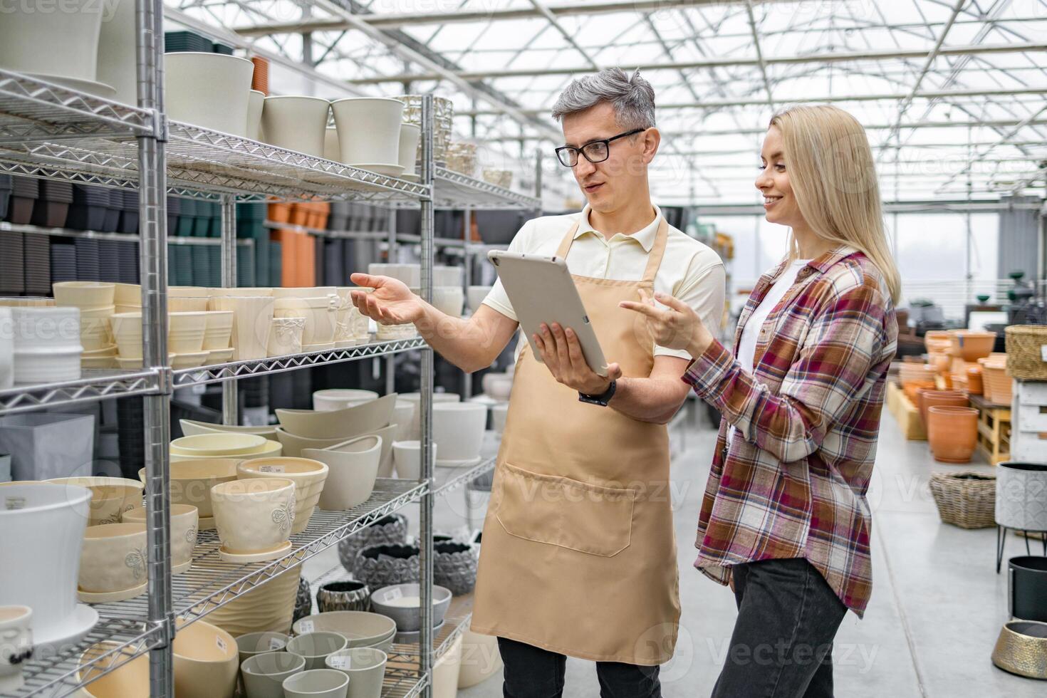 A Caucasian woman discusses flower pots with a shop assistant at a plant store. They're examining options on a tablet device near shelves. photo