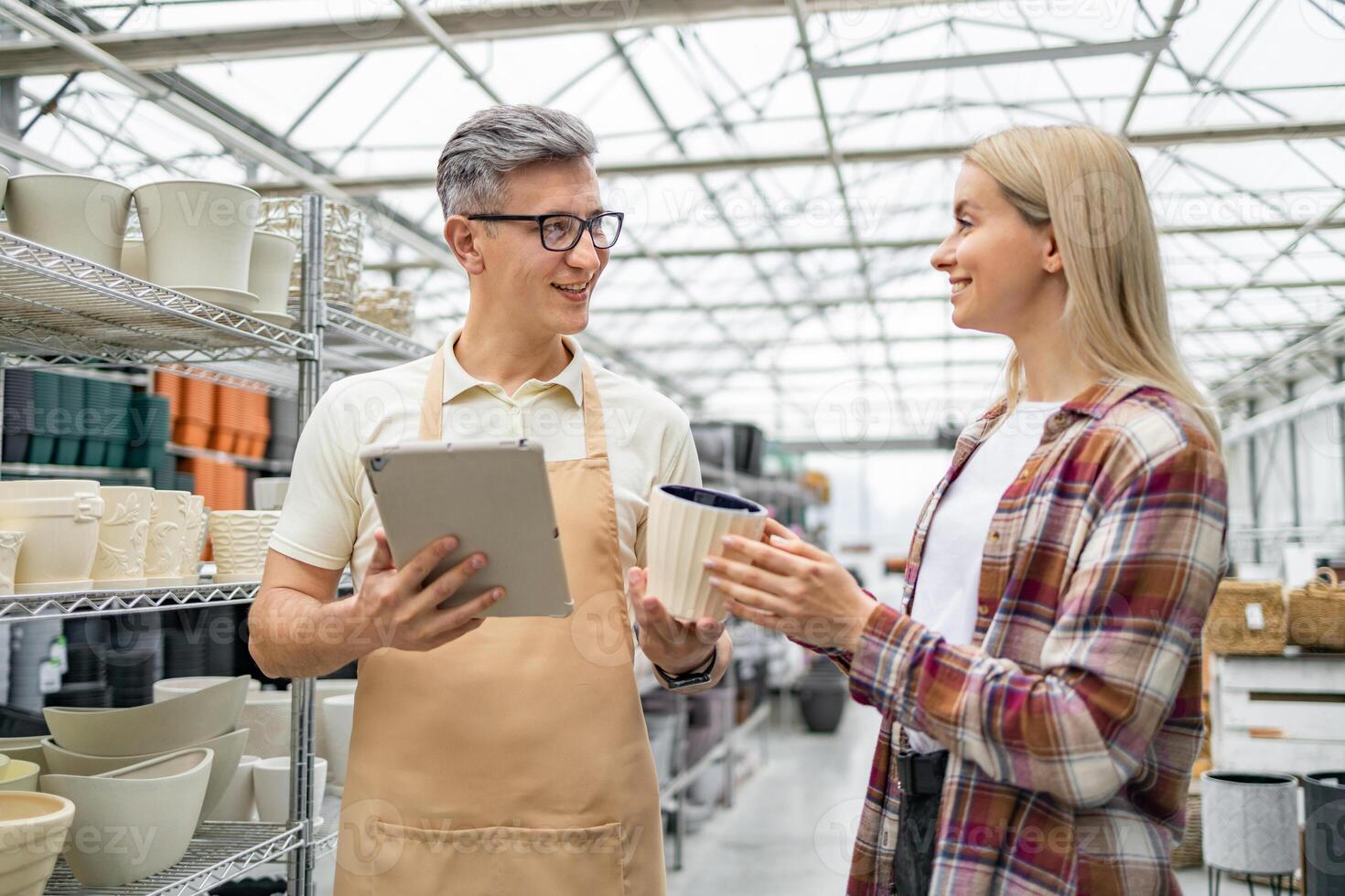 A shop assistant assists a customer in selecting a flowerpot, offering help with a tablet. The interior showcases an array of stylish pots. photo