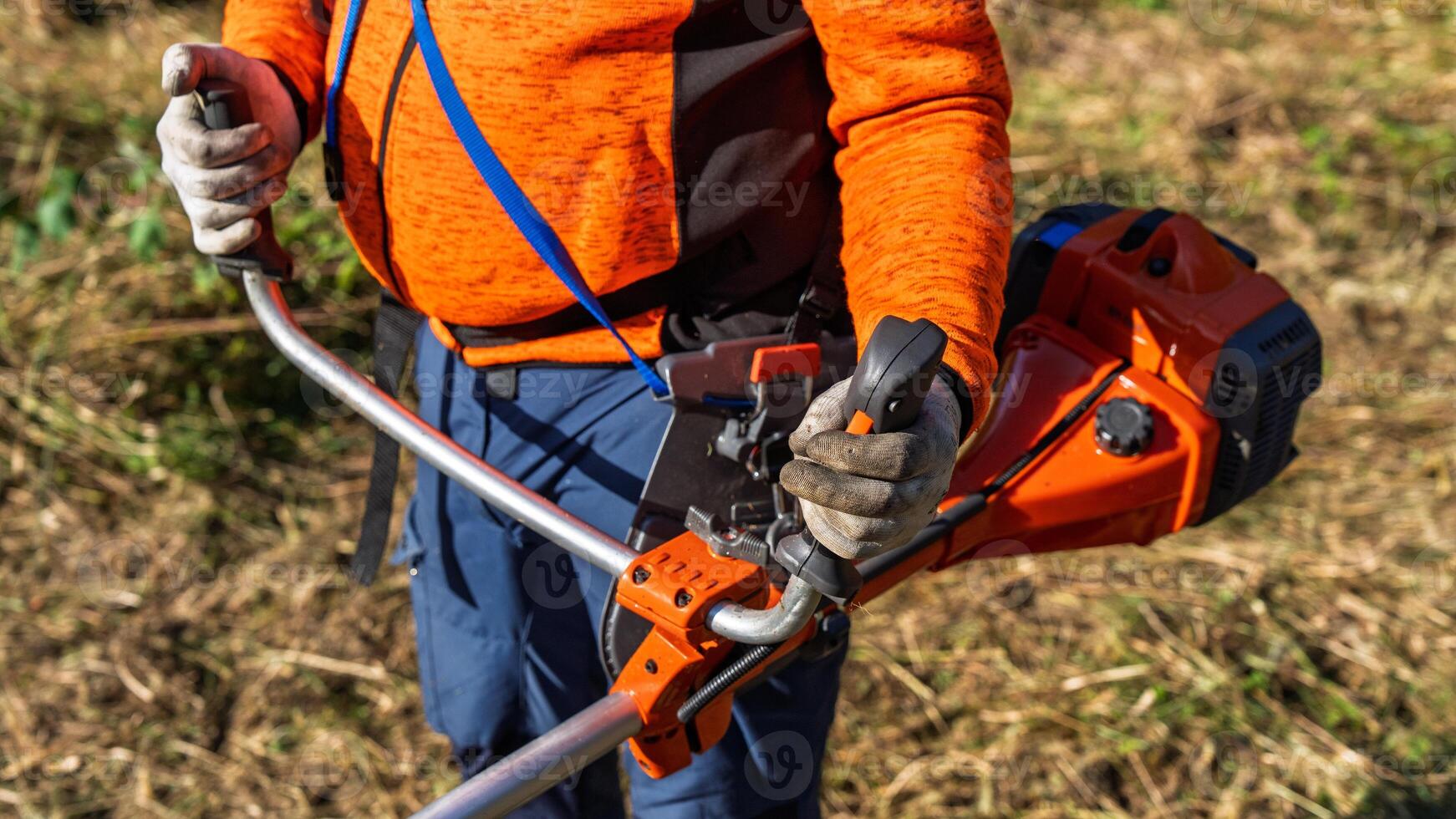 A person using a string trimmer to cut grass in a yard. Wearing gloves and an orange shirt, they focus on lawn maintenance, representing landscaping and gardening tasks. photo