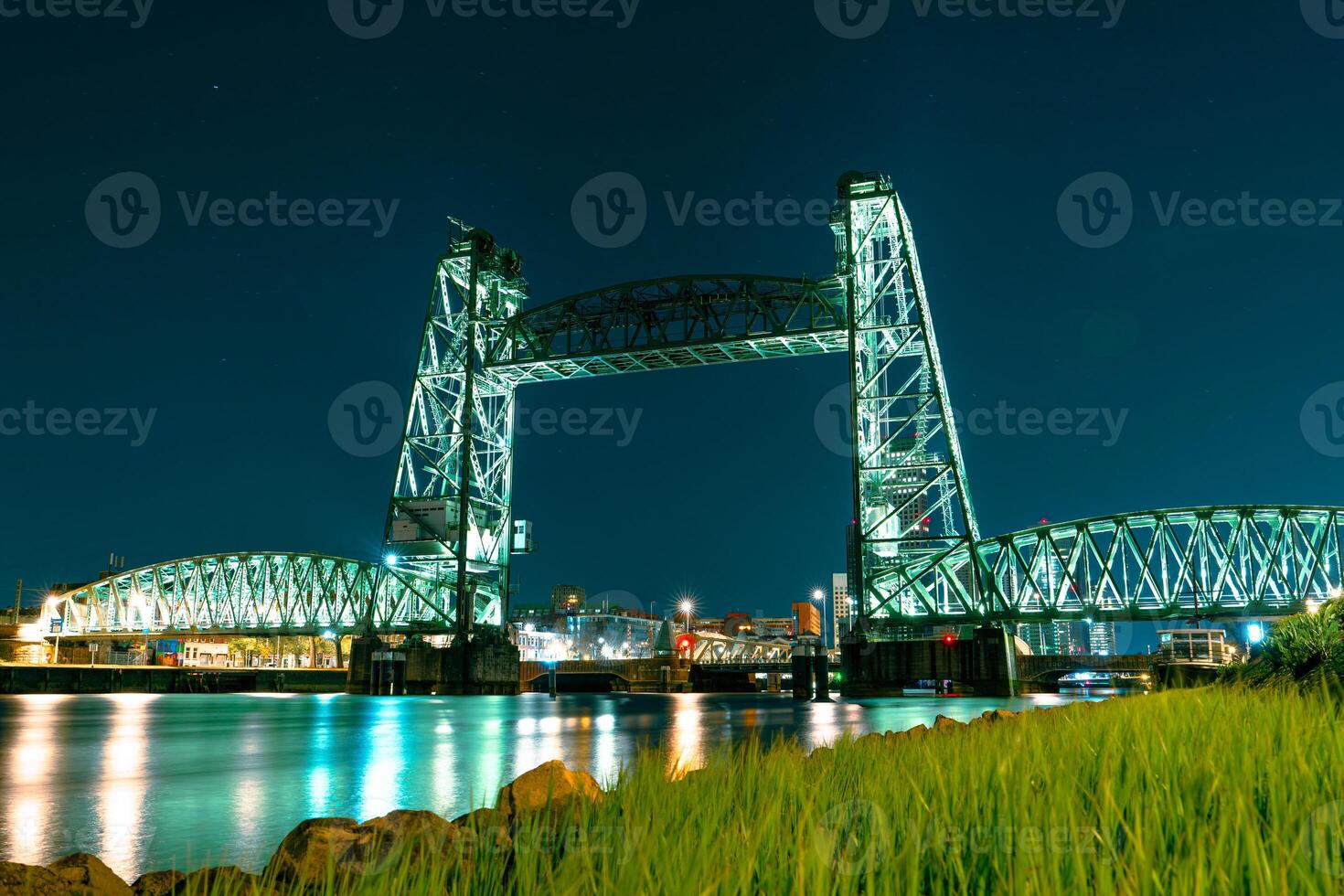A captivating long exposure night photograph of the Koninginnebrug bridge in Rotterdam, showcasing the illuminated urban city skyline and mesmerizing light trails streaking across the waterway. photo
