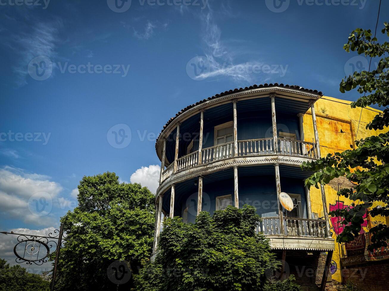 Rustic Georgian balcony house with curved wooden facade and satellite dish, framed by vibrant greenery and dramatic sky in an old historic neighborhood photo