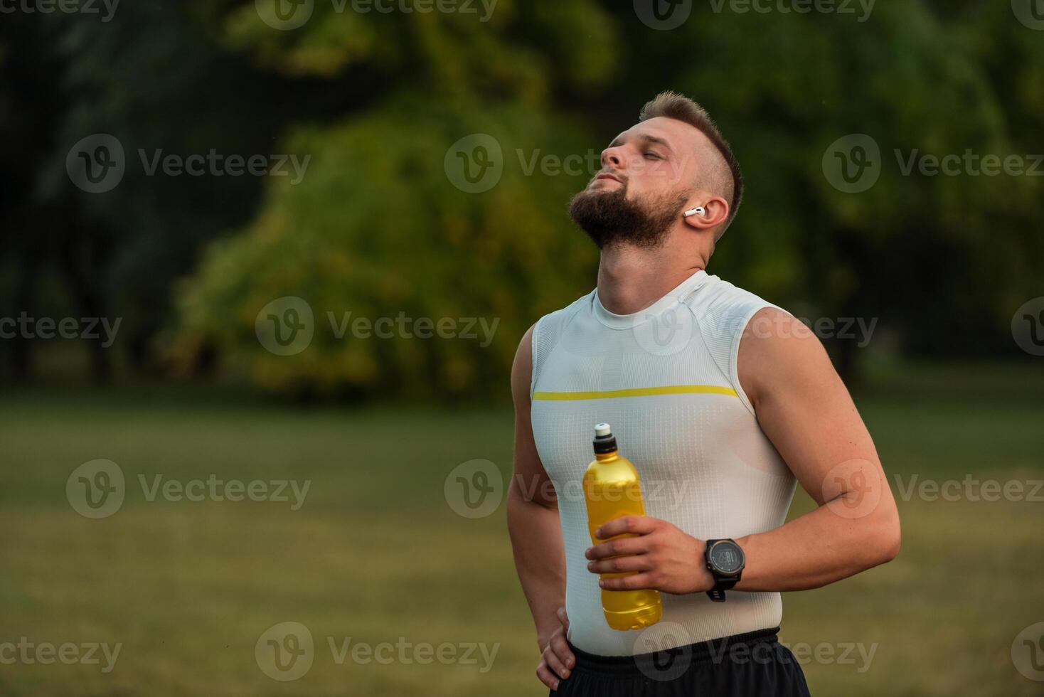 A young fit man with eyes closed listening to music on wireless headphones and relaxing after a run or workout in the park, active healthy lifestyle. photo