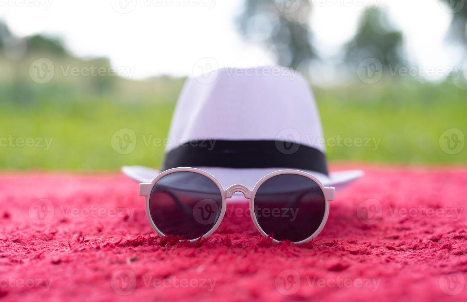 A fashionable white hat with a black band and round sunglasses placed on a red textured blanket. Summer style, vacation mood, and trendy accessories for travel, fashion, and lifestyle concepts. photo