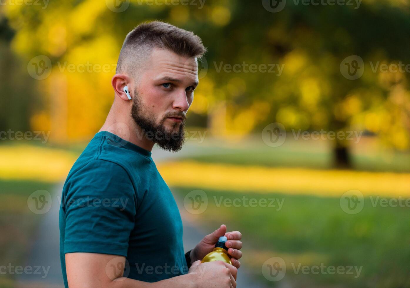 A fit young man listening to music on wireless headphones and drinking water or nutrition juice from a bottle after a run or workout in a summer park active healthy lifestyle.. photo