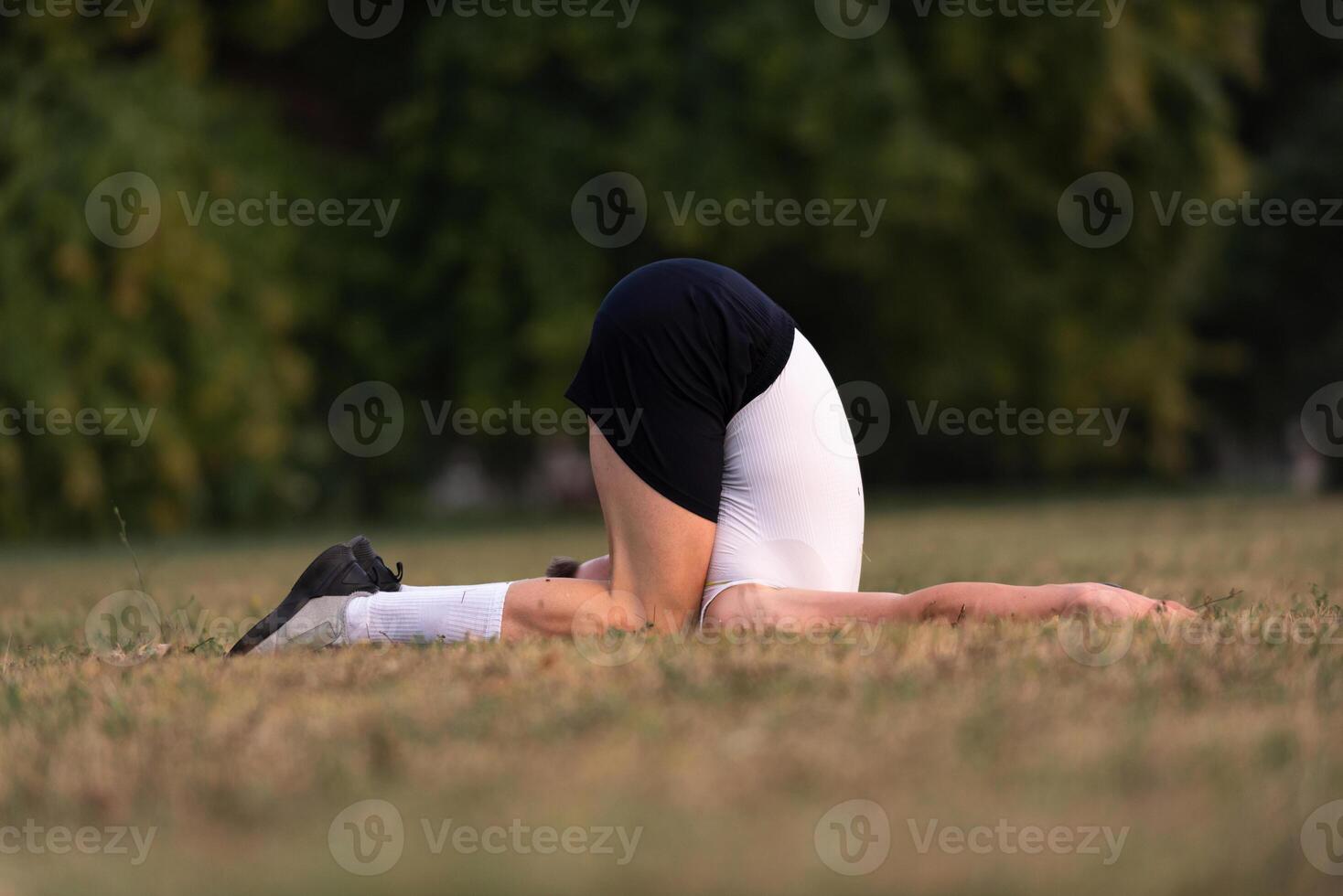 A fit man lying on the grass in a yoga or stretching pose, with head tucked under and legs bent, in a natural park setting. photo