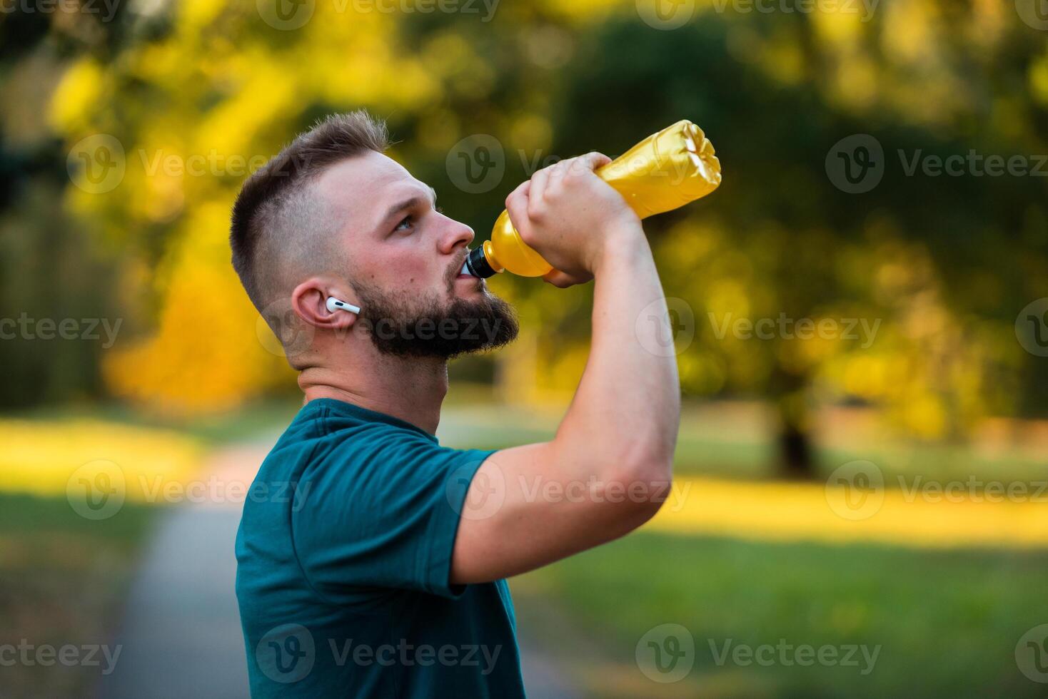 A fit young man listening to music on wireless headphones and drinking water or nutrition juice from a bottle after a run or workout in a summer park active healthy lifestyle. photo