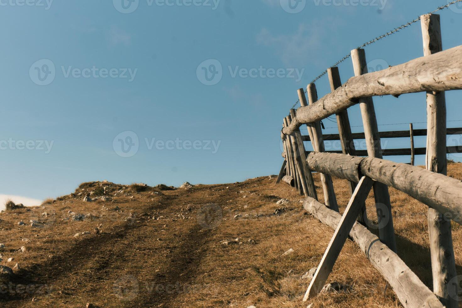 Rural landscape with a wooden fence on a hilly terrain under a clear blue sky. Peaceful natural scene captured in warm afternoon light. photo
