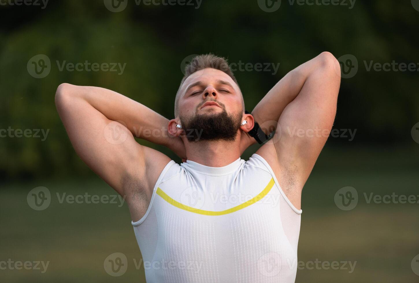 A young fit man stretch before running or workout in a park, he listens to music on wireless headphones with eyes closed to stay motivated. Active lifestyle, fitness and outdoor exercise concept. photo