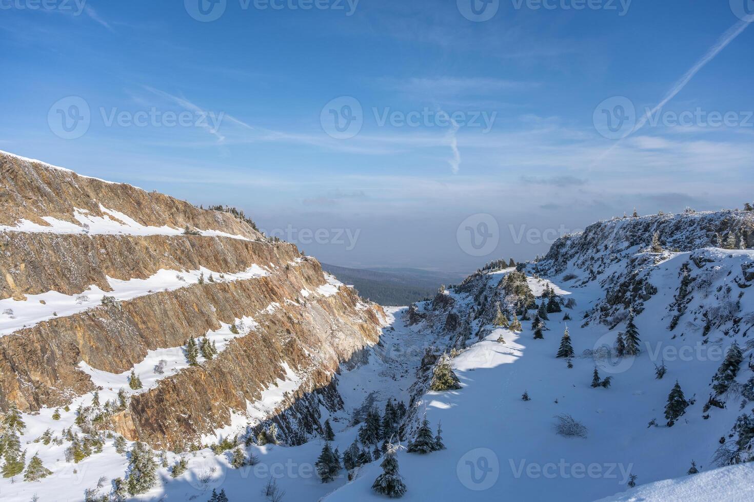 Snow blankets the Stanislaw Quartz Mine in the Izerskie Garby region, creating a serene winter landscape. The rugged cliffs and scattered trees stand out against the clear blue sky. photo