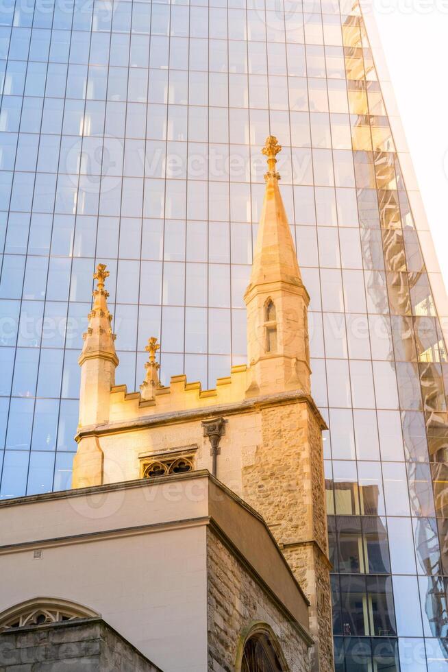 St Andrew Undershaft Church stands in striking contrast to the sleek, glass facade of a modern skyscraper in London. This scene captures the blend of history and contemporary architecture. photo