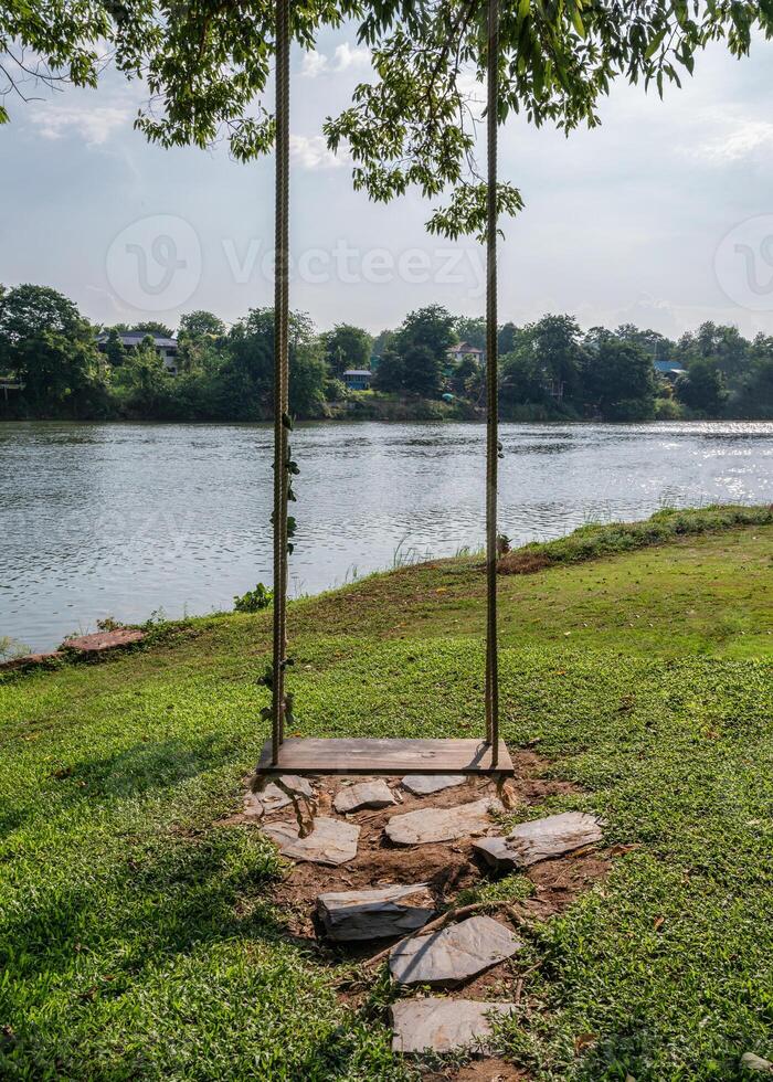 Wooden swing with rope hanging on tree at waterside photo