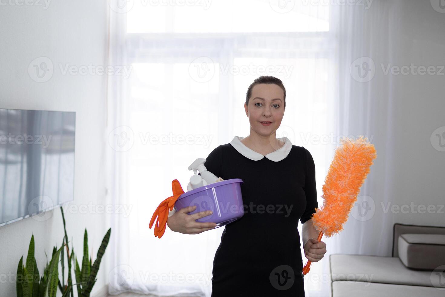 A female housekeeper stands in her modern living room with various cleaning products photo