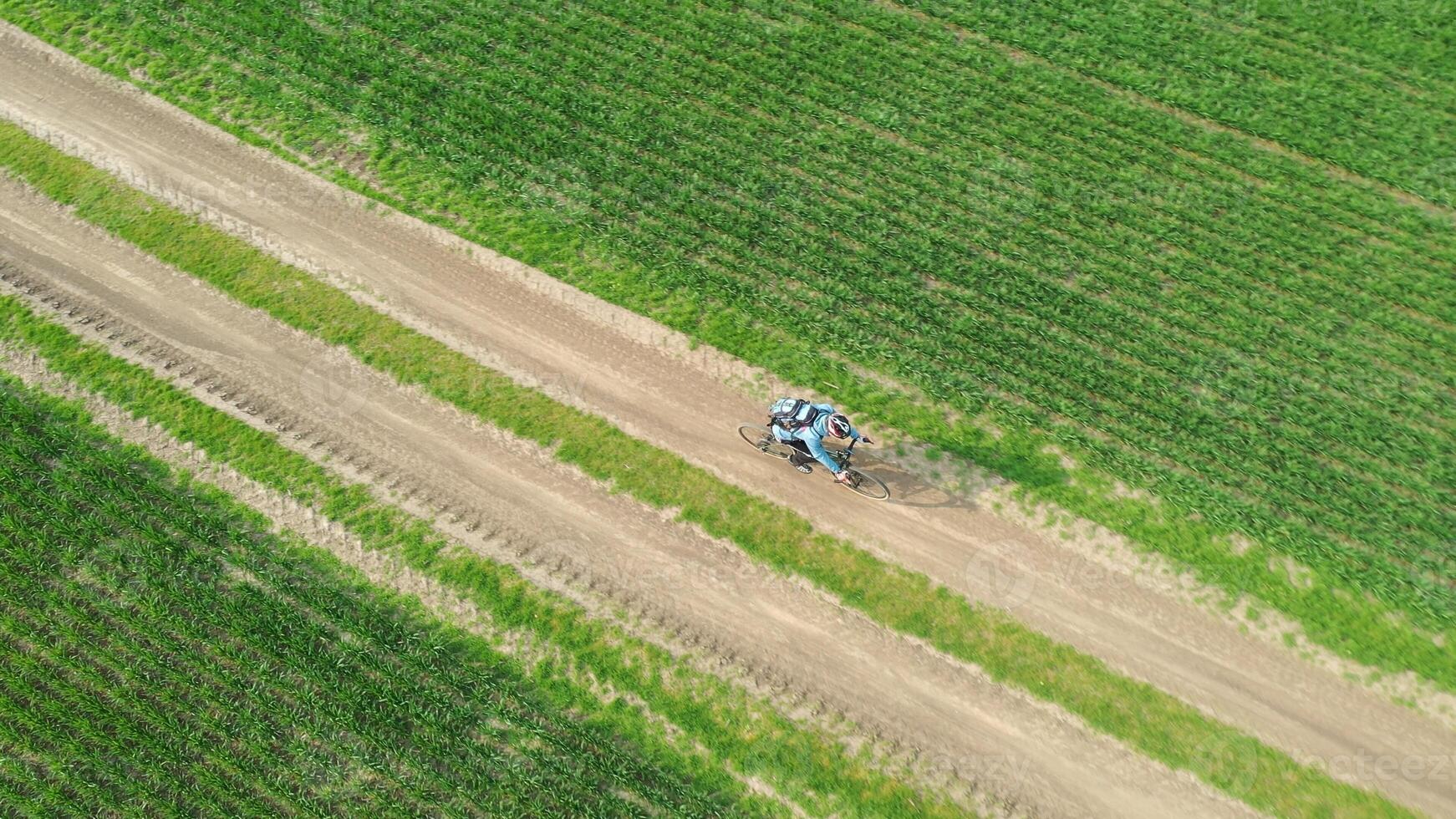 Aerial view of a cyclist on a bicycle riding on a country road between green fields. Cycling traveler riding across the field on a dirt road. photo
