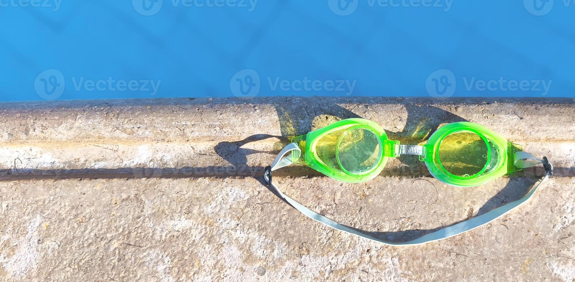 Bright green swimming goggles rest on the edge of a pool, casting a smile-shaped shadow in the sunlight. Perfect summer vibe and vacation mood photo