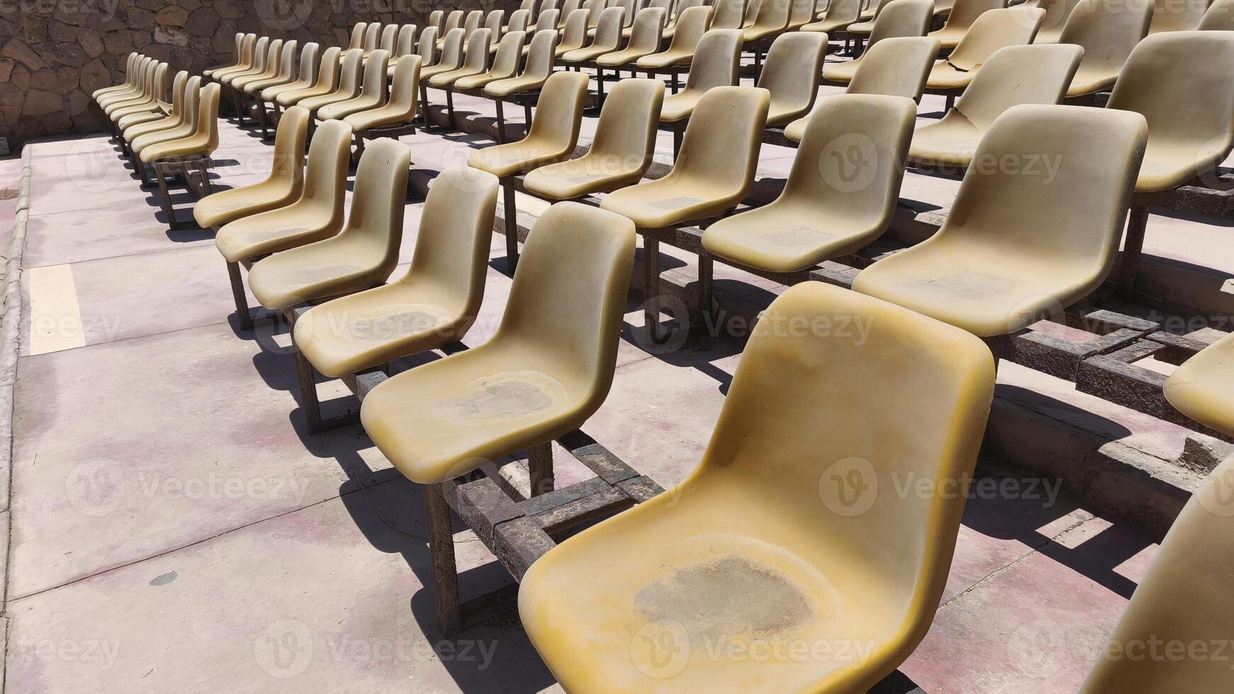 Rows of old, sun-faded plastic stadium seats in an outdoor arena under bright sunlight. The scene evokes a sense of summer heat and empty anticipation before an event photo