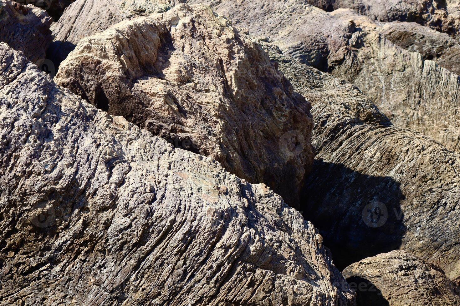 Textured coastal rocks with deep grooves and layers, shaped by centuries of wind and water erosion. The rugged surface contrasts with the soft shadows and sunlight. photo