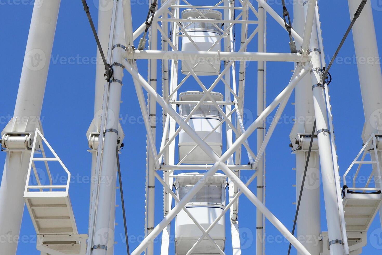 Low angle view of a white Ferris wheel structure with geometric metal supports and cabins, set against a vivid blue sky. The image captures architectural symmetry and summer leisure. photo