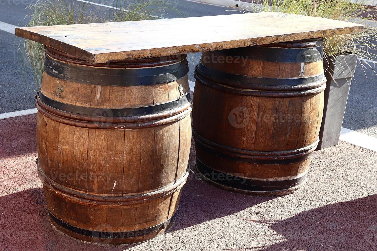 Two rustic wooden barrels with black metal bands support a thick wooden tabletop, forming a unique outdoor table setup on a red pavement, blending function and vintage style. photo