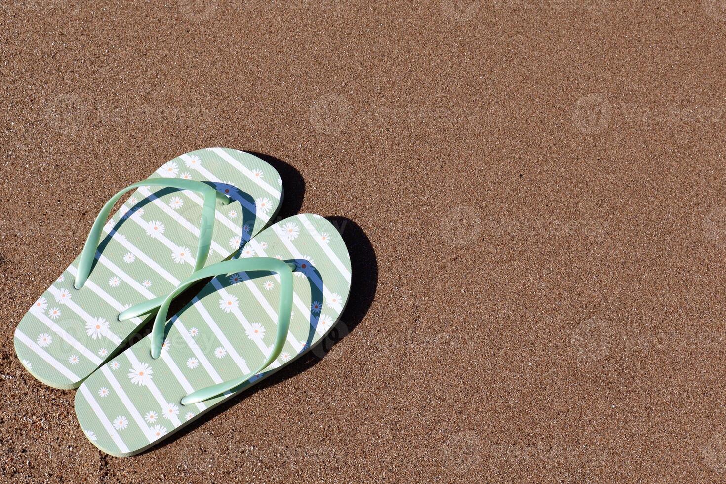 A pair of green flip-flops with white daisy patterns lies on warm golden sand. The image captures a peaceful summer vibe, perfect for vacation or travel themes. photo