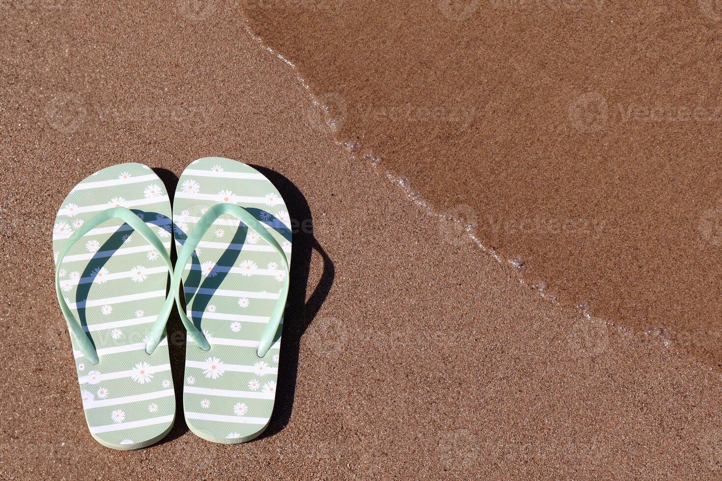 A pair of light green flip-flops with a daisy pattern rests on the wet sandy beach as a gentle wave approaches. The photo captures the essence of a relaxing summer day.