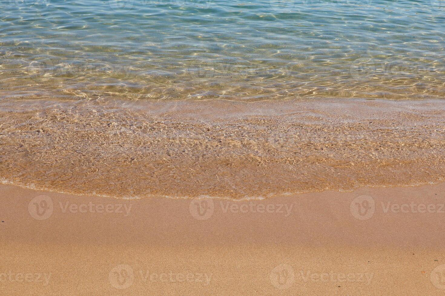 A gentle wave rolls onto a sandy beach under clear skies. The water is crystal-clear with shades of turquoise and blue, creating a peaceful and relaxing summer scene. photo