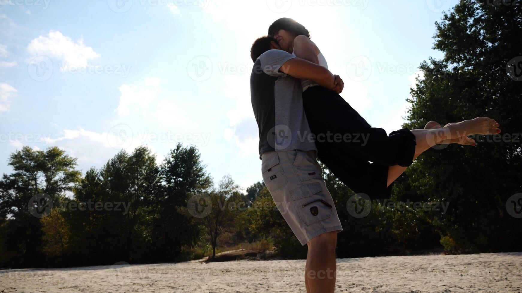 Happy couple having fun together on seashore at summer vacation.Man taking woman in air and turning around at coastline. Carefree lovers spending time at sandy beach. Concept of love. Slow motion photo
