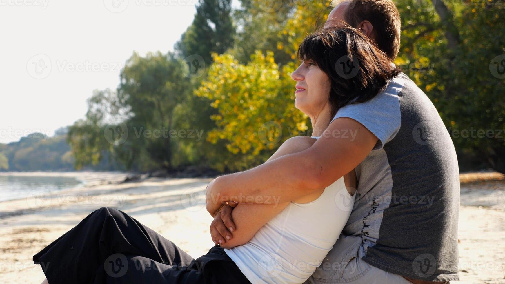 Happy pair hugs during siting on wooden log on beach. Romantic couple admiring sea landscape at sunny day. Lovers spending time together at resort. Concept of summer vacation or holiday. Slow mo photo