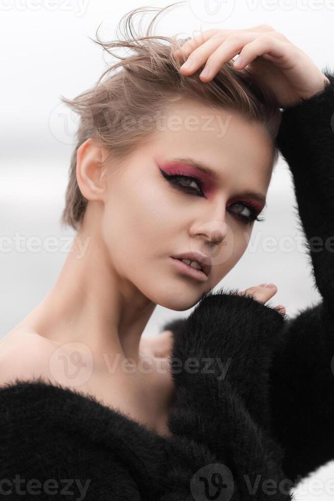 Portrait gorgeous blonde woman is epitome of sensuality. She is wearing short hairdo and bright makeup and is posed outdoors against white natural background, showcasing her beauty in all its glory photo