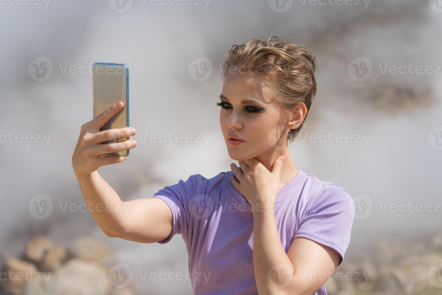 Beautiful female traveler using her cell phone to record self portrait to call with friends in remote location against backdrop of steaming geysers in crater of an active volcano while traveling photo