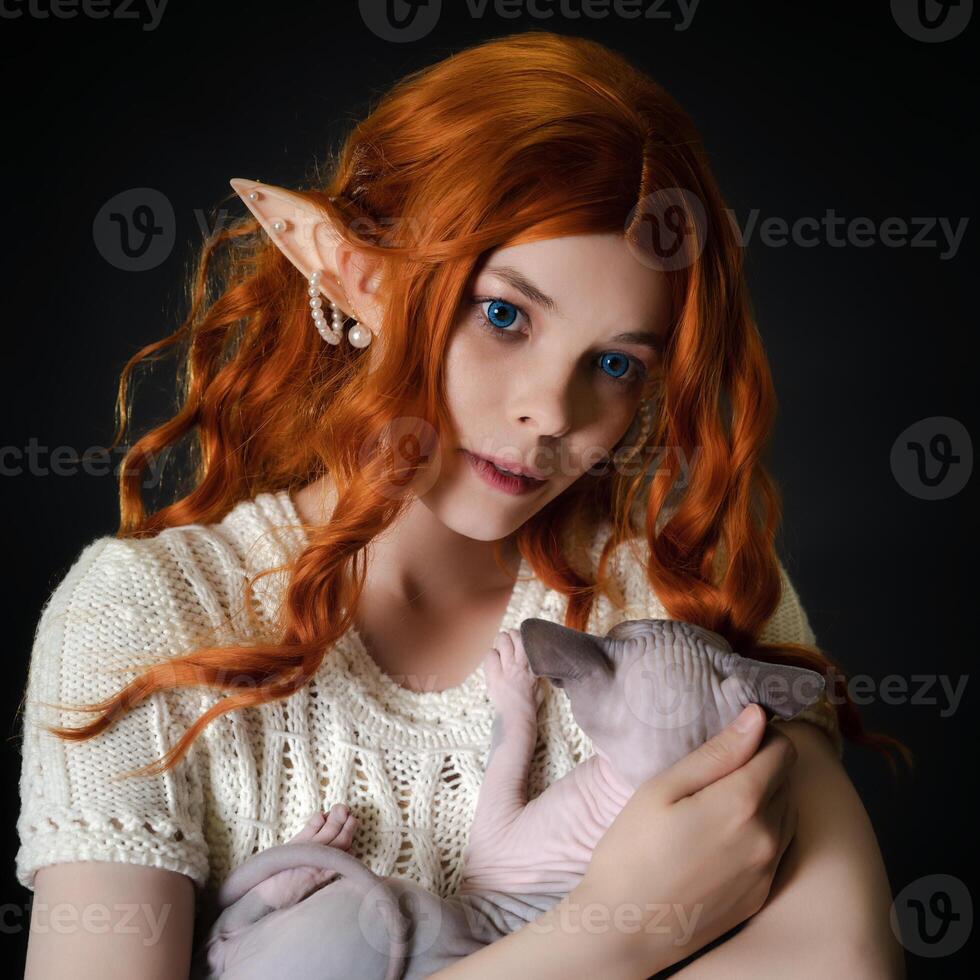 Close-up view of ginger young woman cosplayer elf in white knitted dress, with long wavy hair, blue eyes, cradling Sphinx kitten and looking away. Portrait on black background. Part of Series. photo