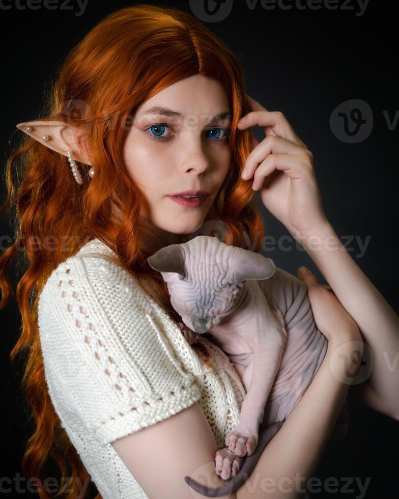 Young female elf cosplay with sleeping Sphinx kitten. Close-up portrait on black background. Cute elf in white knitted dress, with long red curly hair, blue eyes is looking at camera. Part of Series. photo