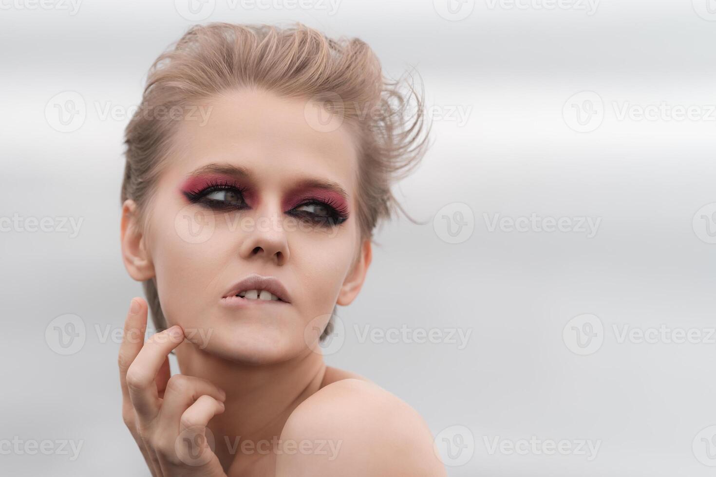 Emotionally stressed woman with bright makeup strikes puzzled pose, pursing her lower lip, raising palm to neck and looking away. Soft selective focus, natural white background and plenty copy space photo