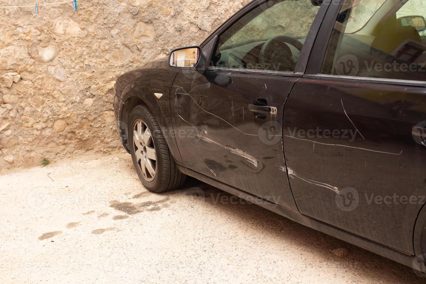 Exploring a neglected black car with scratches parked against a stone wall in a quaint Spanish neighborhood photo