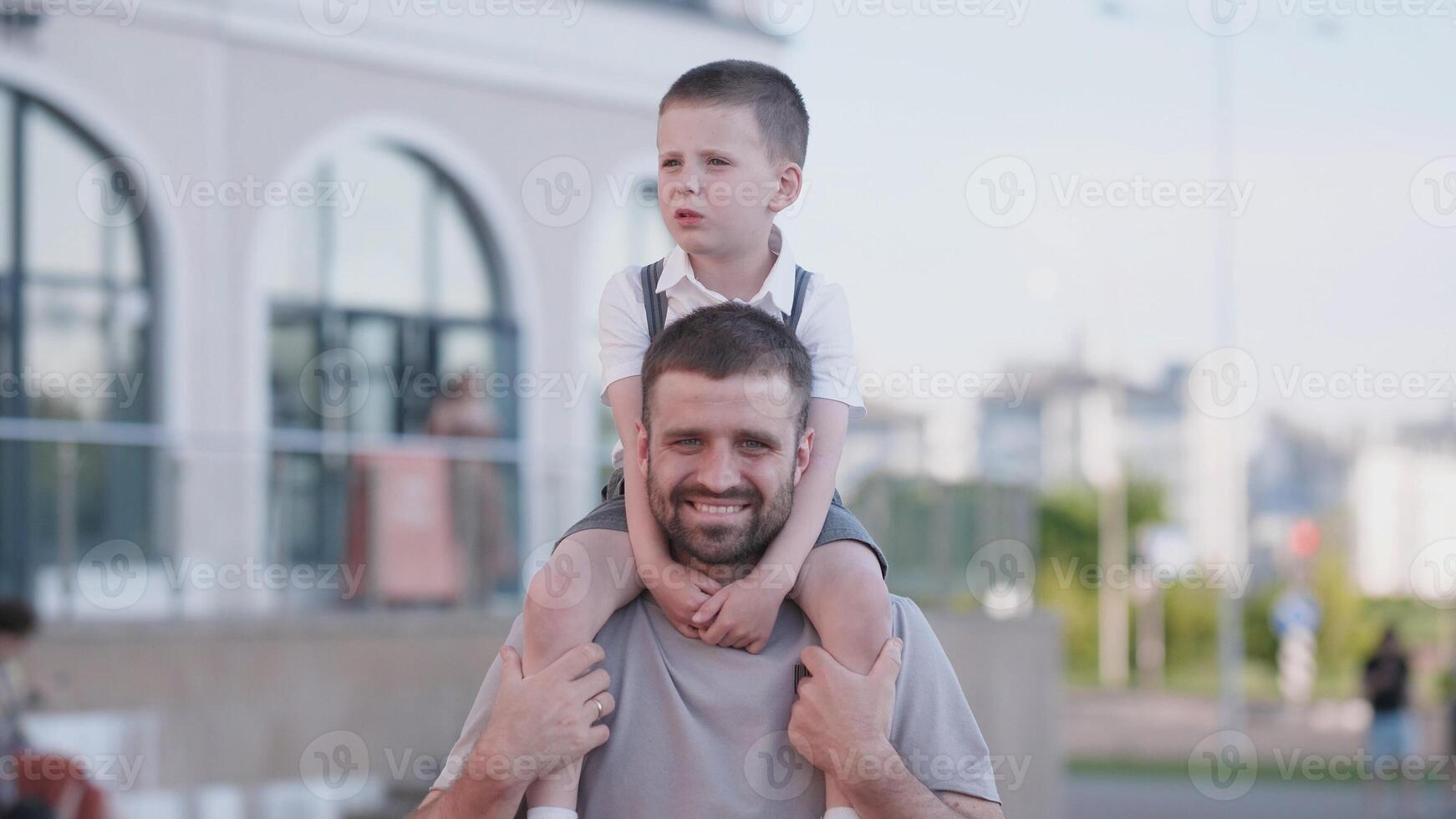 Father and son share joyful moments at a city park, with the child sitting on the father's shoulders, both smiling under the bright sun, surrounded by greenery. photo