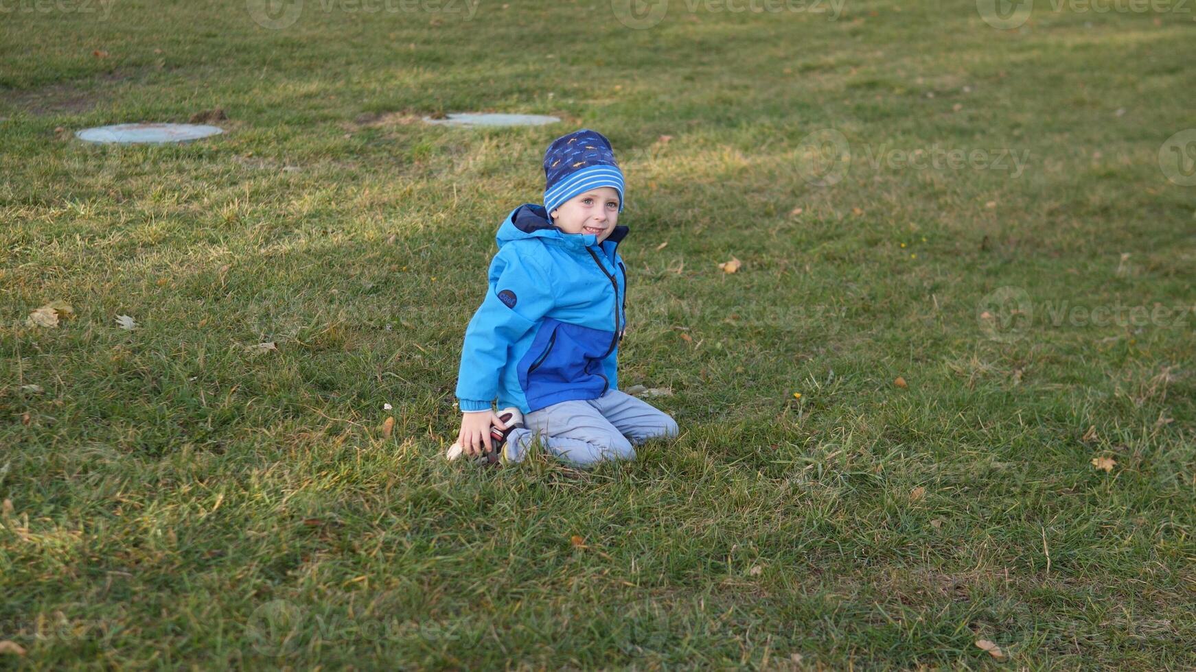Brightly dressed child sits on the grassy ground in the park, smiling and enjoying a carefree moment under the clear autumn sky, surrounded by nature's calmness. photo