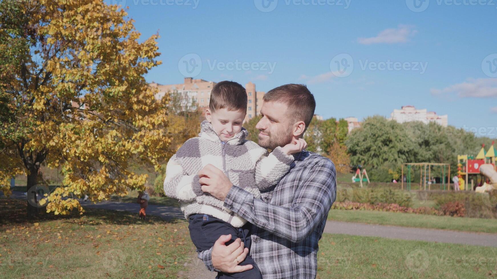 In a bright autumn park, a father gently holds his son, surrounded by colorful foliage and a playground, enjoying quality time on a sunny day. photo