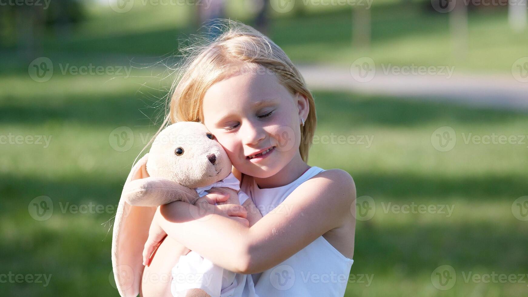 A girl with long blonde hair hugs a soft bunny toy while standing in a bright, green park, surrounded by trees and enjoying a sunny afternoon. photo
