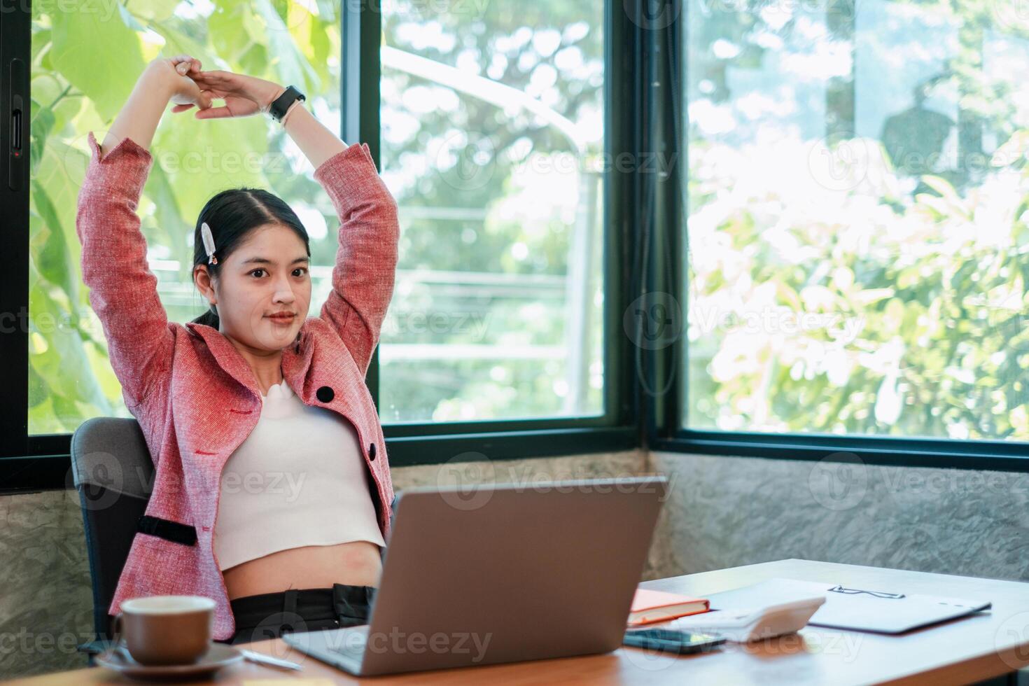 A young woman in a pink blazer stretches at her desk in a bright, modern office setting with a laptop and coffee. photo