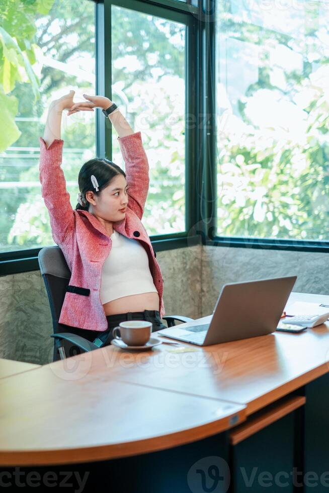 A young woman in a pink jacket stretches at her office desk, with a laptop and coffee, in a bright, modern workspace. photo
