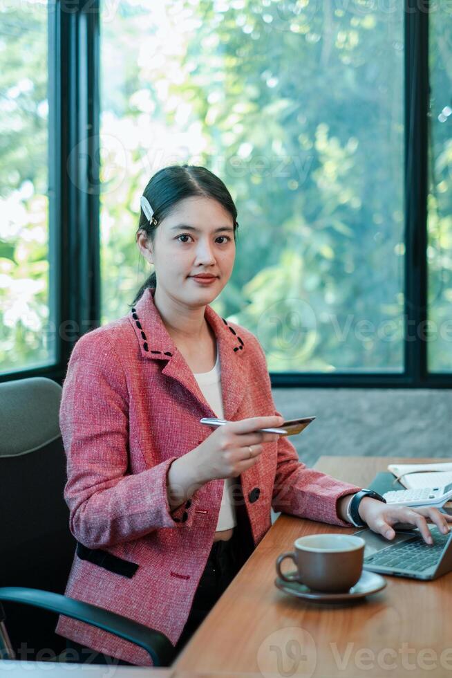 Businesswoman in a pink blazer working at a desk with a laptop and coffee, set against a backdrop of lush greenery through large windows. photo