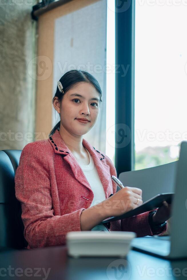 A confident woman in a pink blazer uses a tablet in a bright, modern office environment. photo