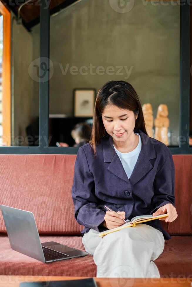 A young woman in a casual setting writes in a notebook with a laptop beside her, capturing a moment of focus and creativity. photo