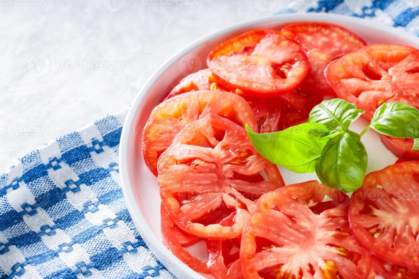 Tomato Slices with Basil Leaves on White Plate, Blue Checkered Napkin, Light Grey Background, Top View photo
