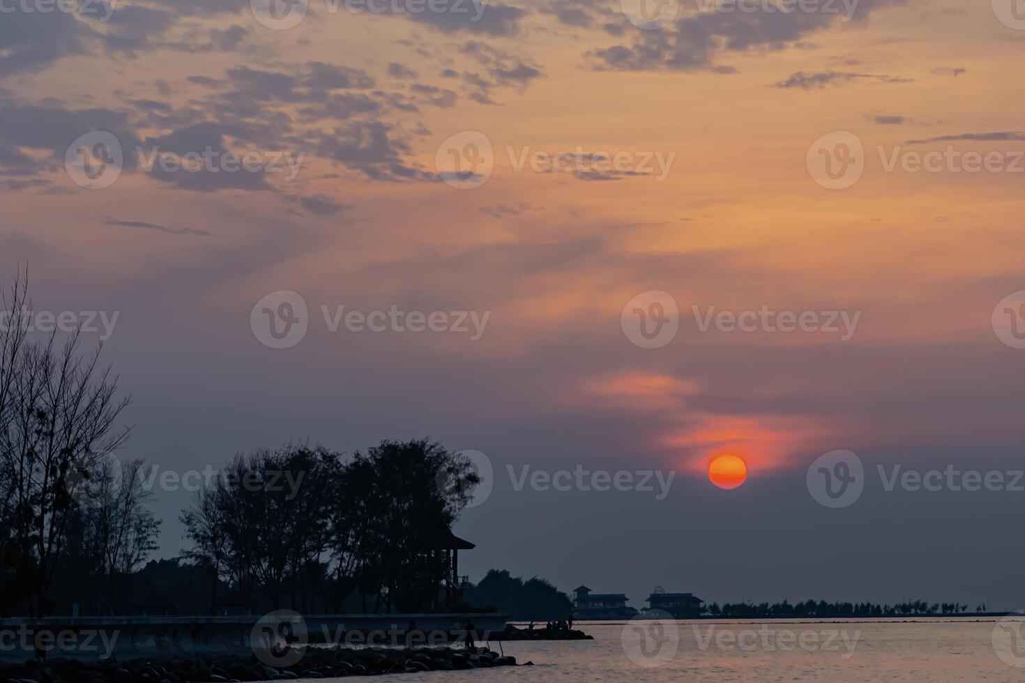 Beautiful sunset over the sea with glowing orange sky and silhouettes of trees and houses. Calm and dreamy coastal photo
