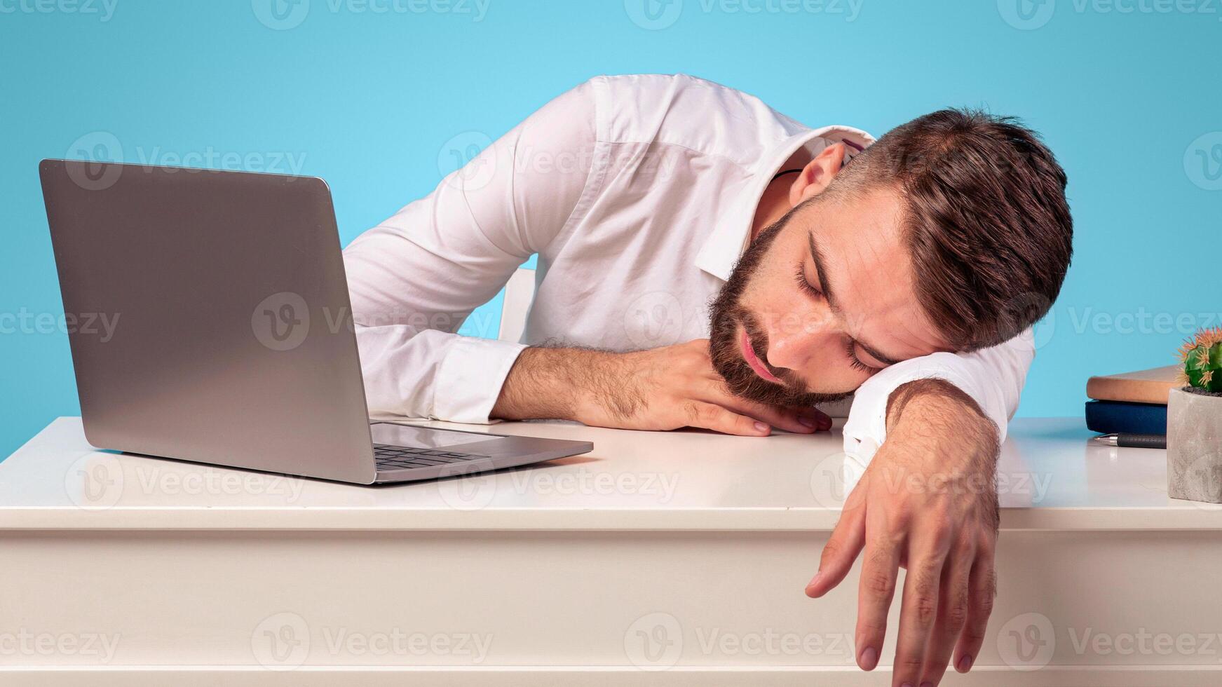 Exhausted young businessman sleeping on his desk, tired of overworking on blue studio background, copy space. Millennial workaholic suffering from chronic fatigue at office, panorama photo