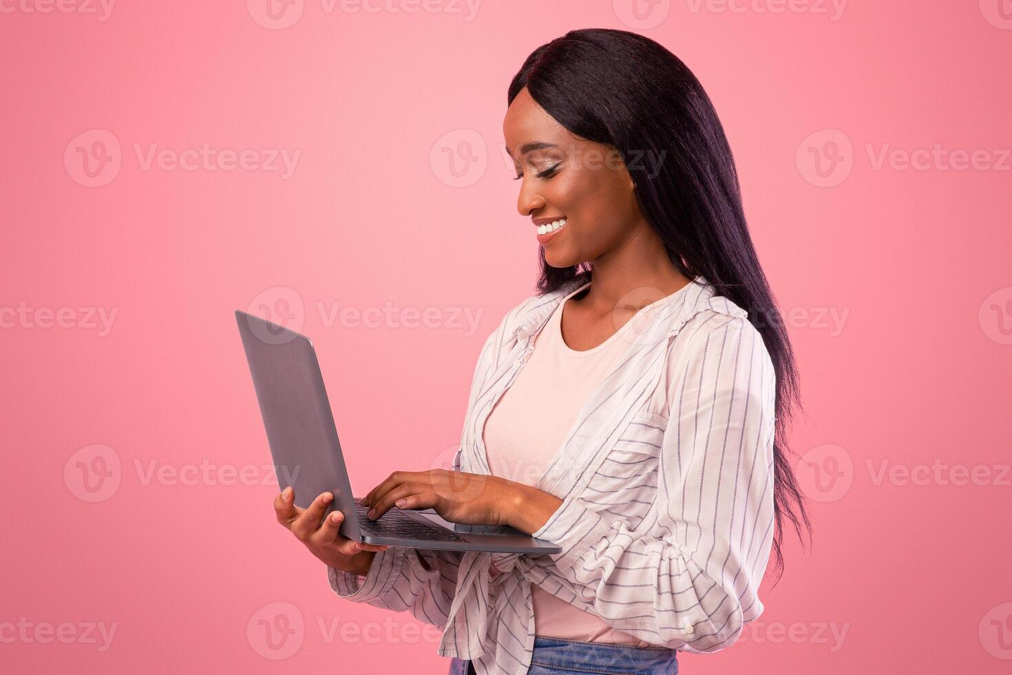 Millennial African American woman working with laptop computer, working or studying online on pink studio background. Lovely black lady communicating on web, watching movie or photo