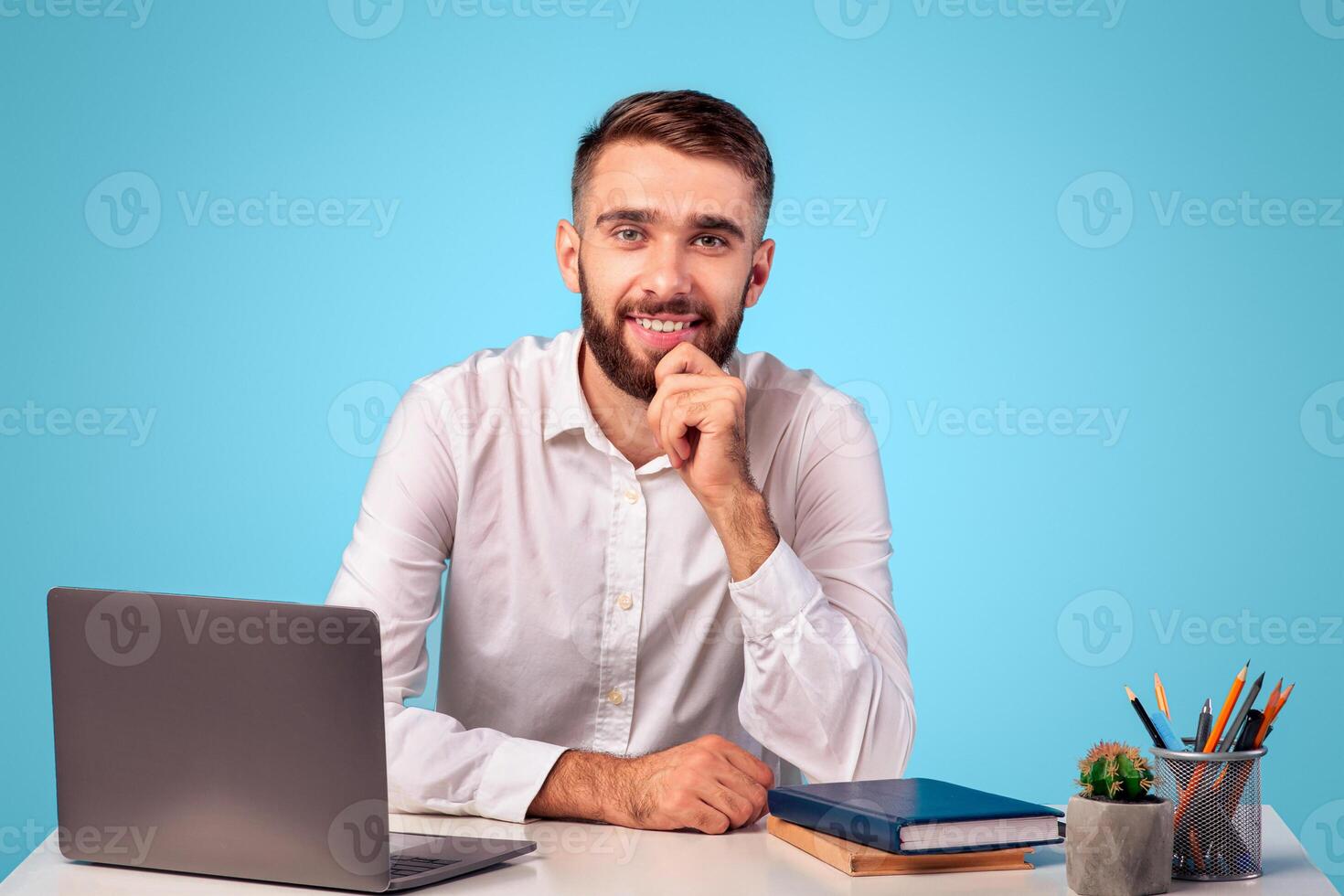 Confident millennial businessman sitting at his desk with laptop computer and looking at camera on blue studio background. Portrait of positive young entrepreneur at workplace photo
