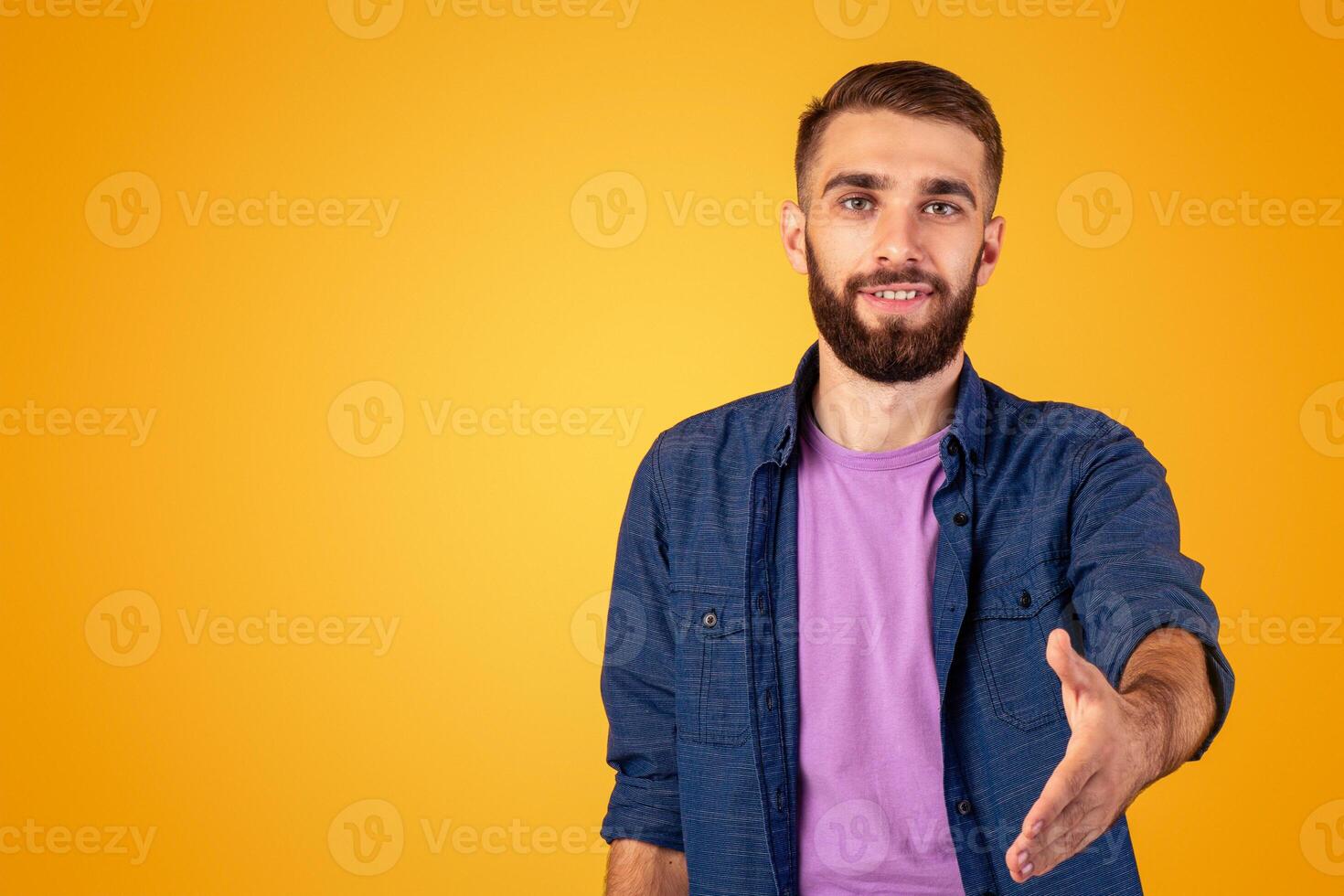 Greeting concept. Smiling Caucasian guy holding out his hand for handshake on orange studio background. Positive young man making agreement or deal, offering partnership or friendship photo