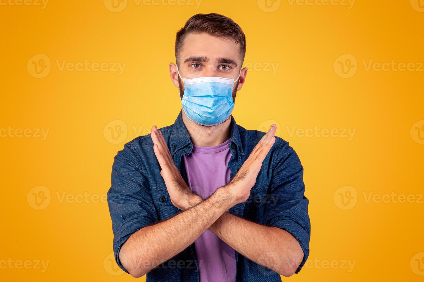 Fighting coronavirus concept. Serious Caucasian man in face mask gesturing STOP on grey studio background. Cool young guy expressing negative attitude to global pandemic, saying NO to virus photo